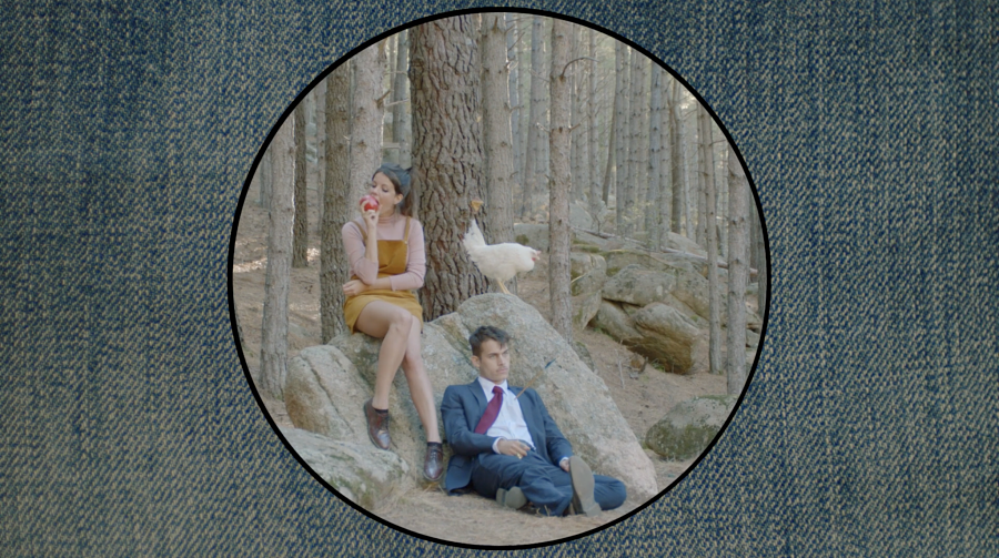 A man and woman sit on rocks with a white bird in a forest, viewed through a circular frame against a denim background.