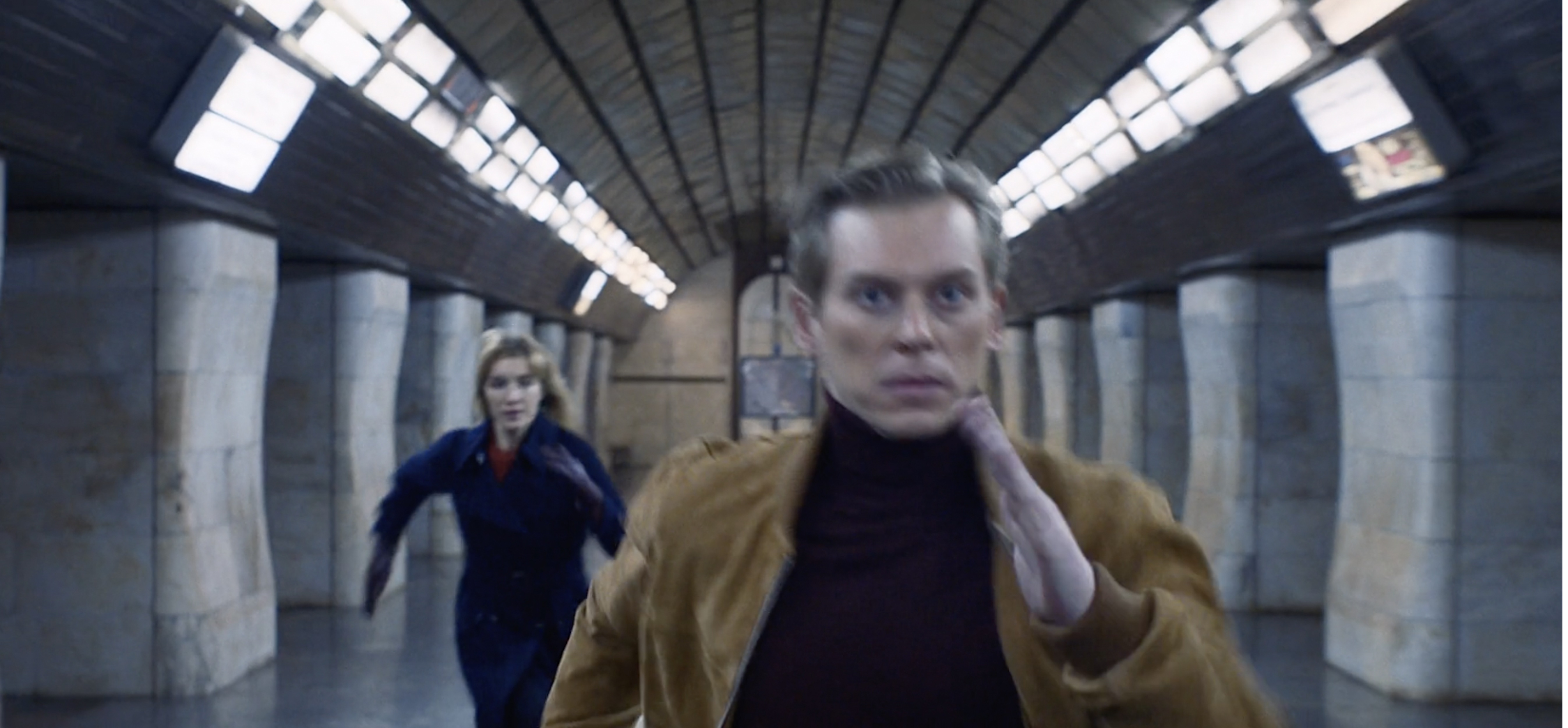 A man and woman are running in a subway station tunnel with concrete pillars, with a shallow depth of field focusing on the man.