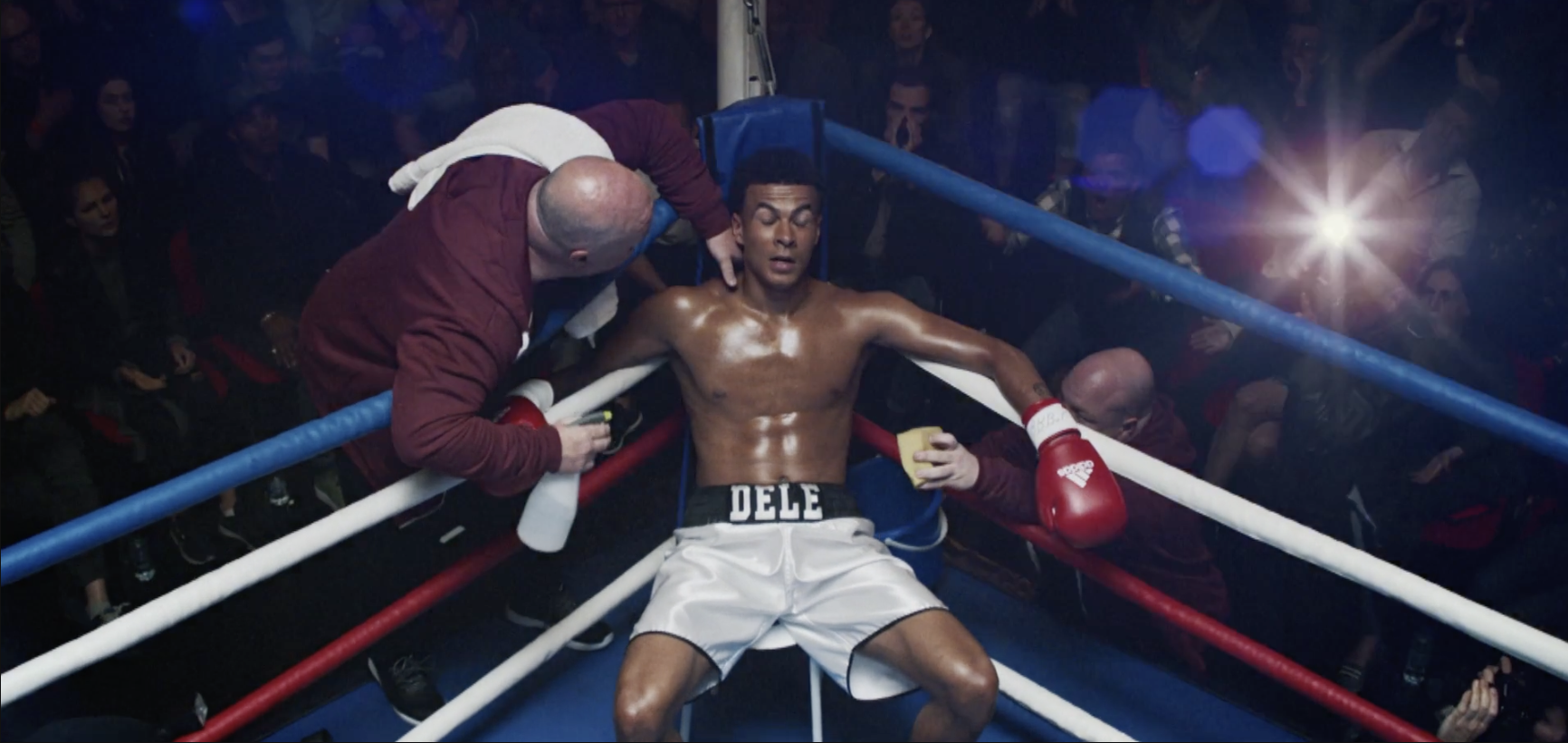 Boxer sits in corner of ring with trainer tending to him. Overhead shot of boxing match with crowd in background, bright lighting.