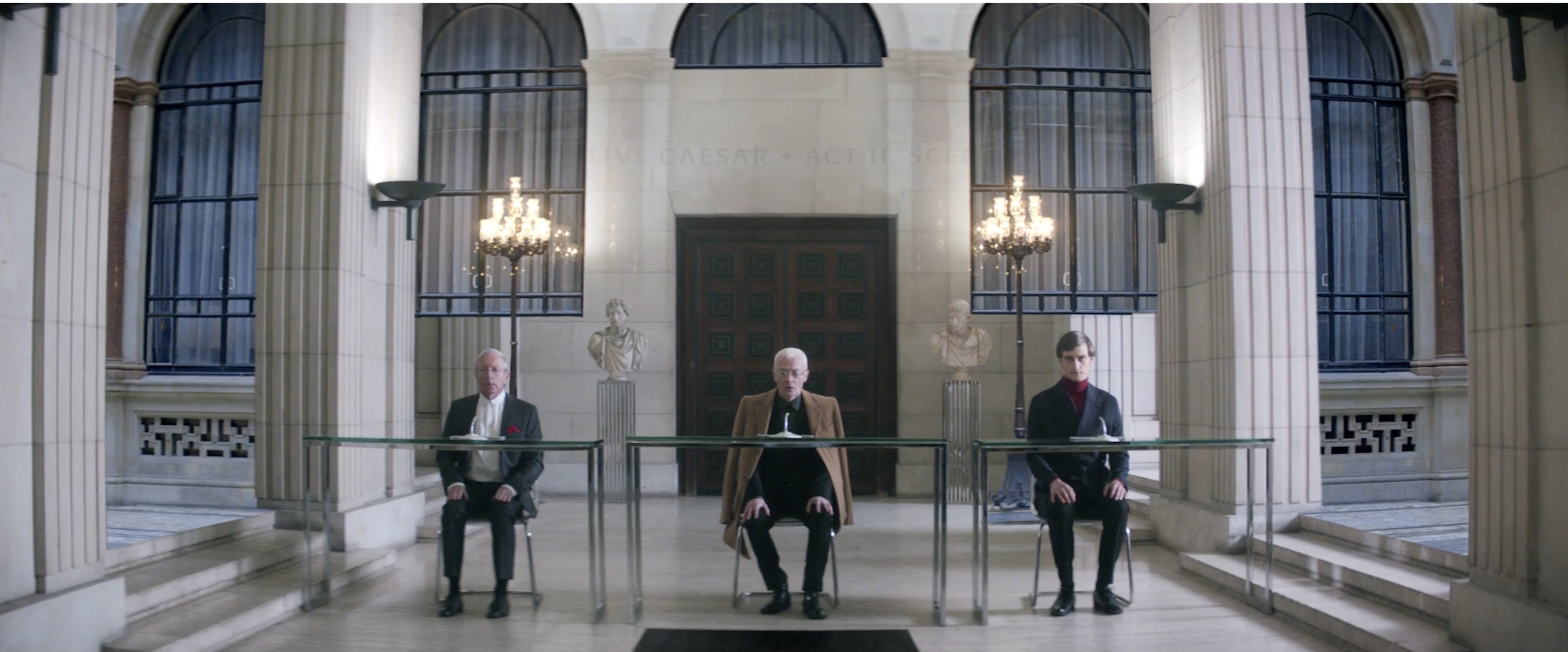 Three men seated at tables in a grand hall with columns, windows, chandeliers, and busts, creating a formal, symmetrical composition.