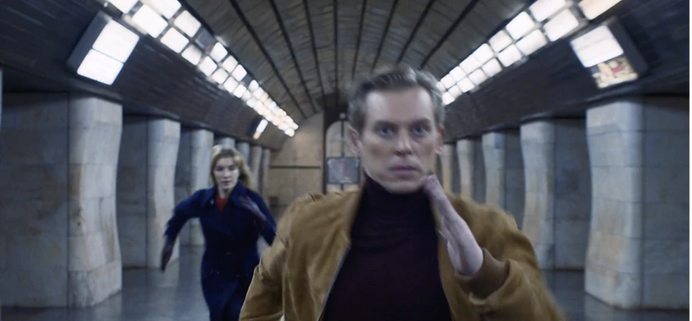 Man in a tan suede jacket running inside a tiled underground station with a woman following behind him.