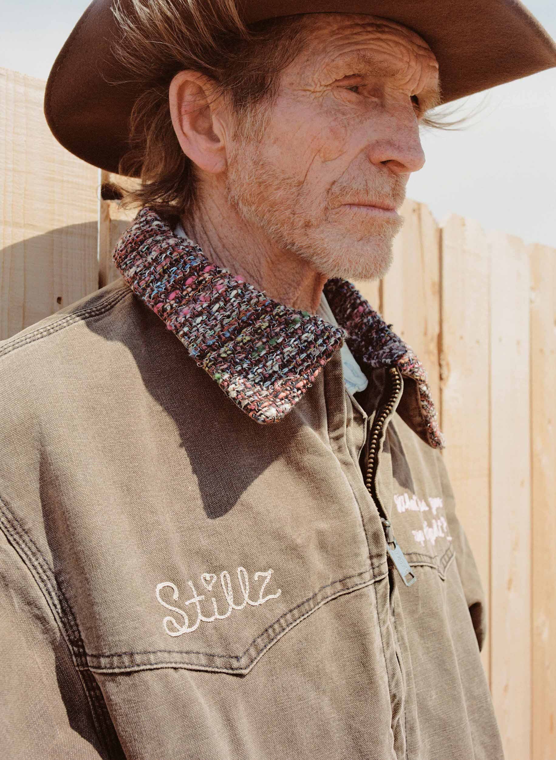 Elderly man in cowboy hat and embroidered jacket stands outdoors by a wooden fence, close up portrait, in natural daylight.