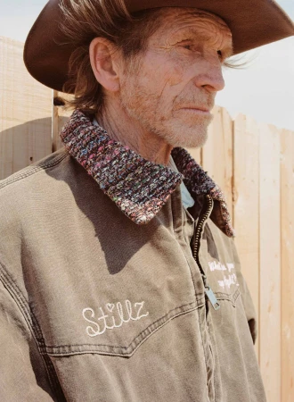 Elderly man in cowboy hat and embroidered jacket stands outdoors by a wooden fence, close up portrait, in natural daylight.