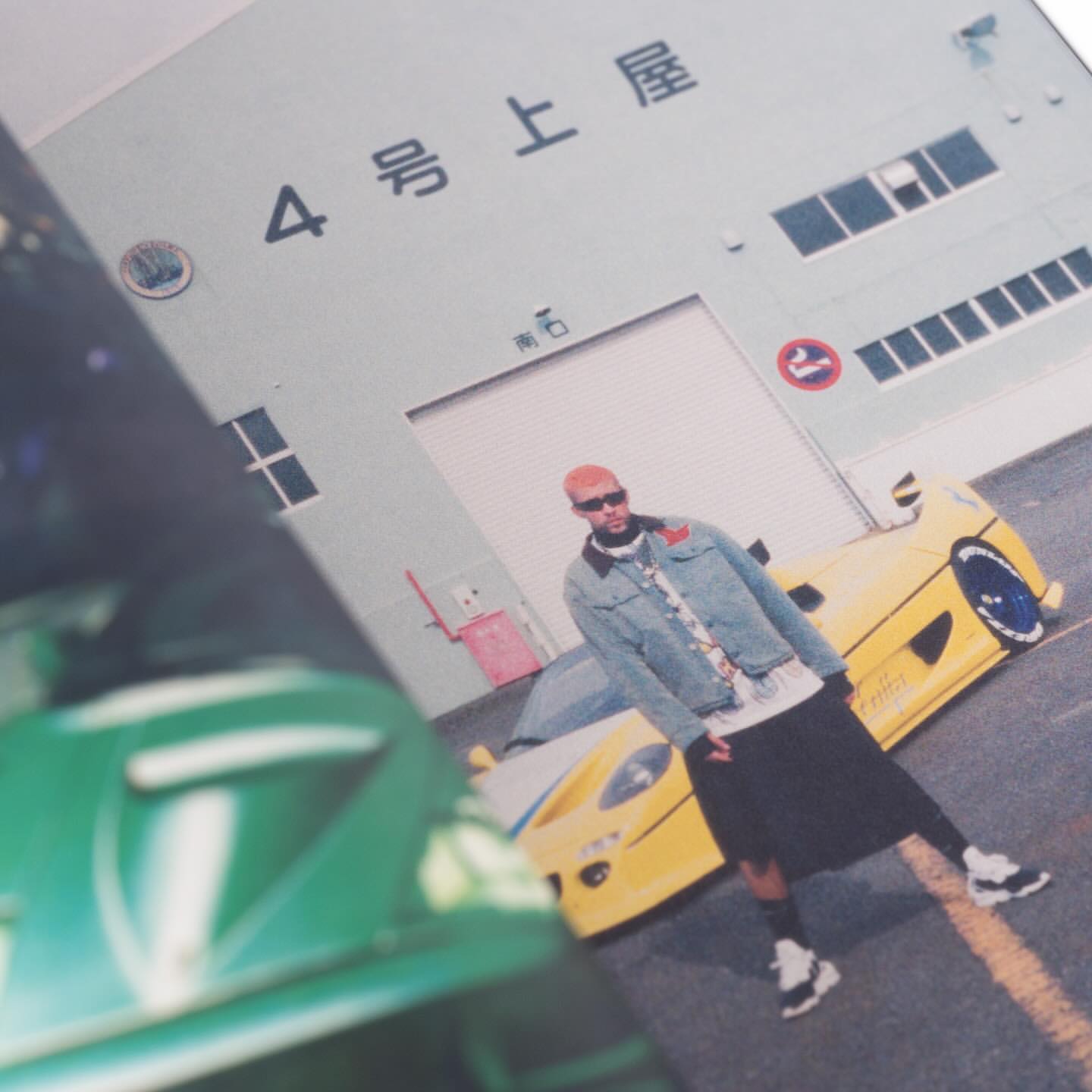 An adult male poses in front of two sports cars, one green and one yellow, with a building in the background, captured at eye-level.