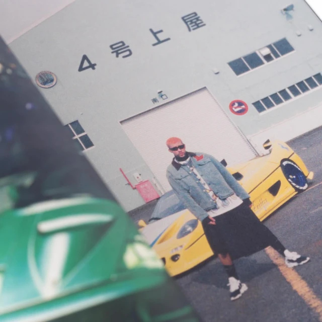 An adult male poses in front of two sports cars, one green and one yellow, with a building in the background, captured at eye-level.