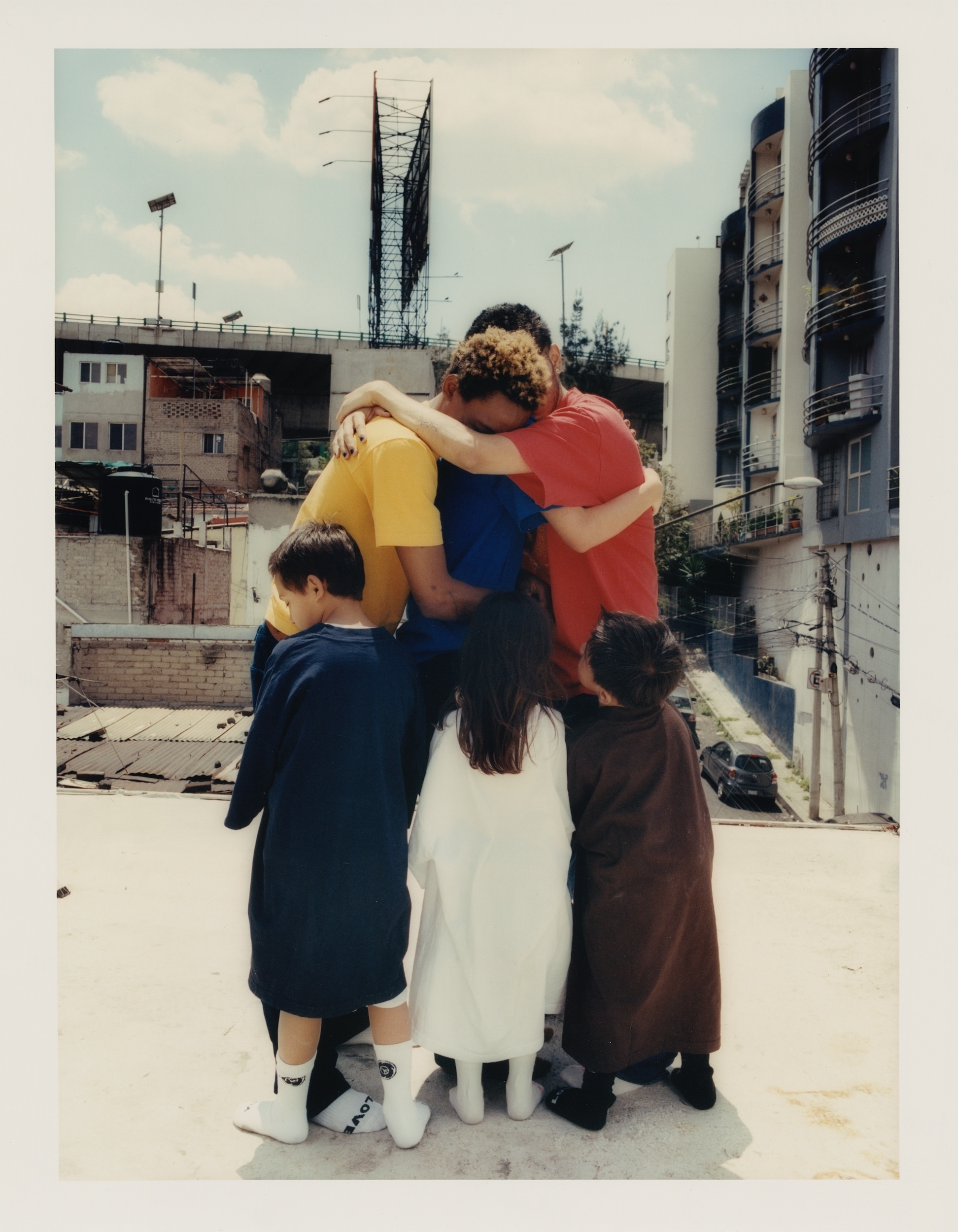 Group of five figures, including three children, embrace outdoors on a rooftop with city buildings in the background during daytime.