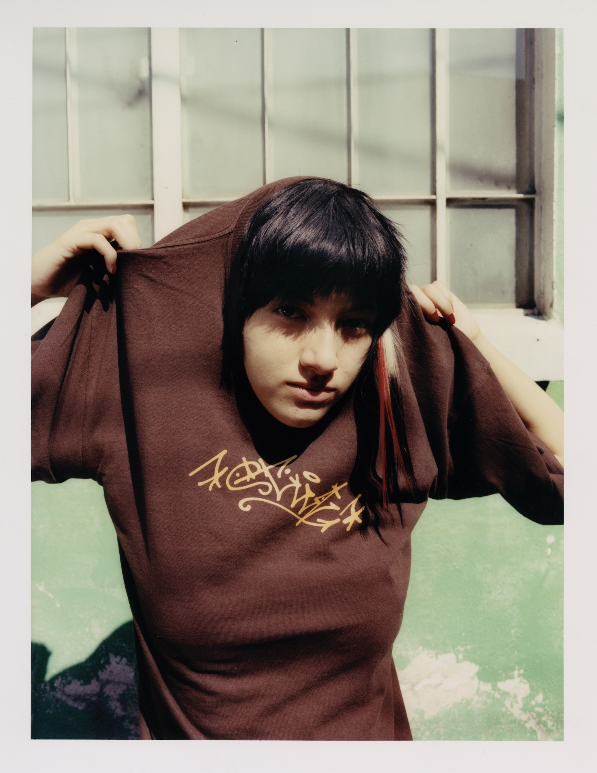 Portrait of a young woman with dark hair and a brown t-shirt with a graffiti logo posing indoors near a window and a light green wall.