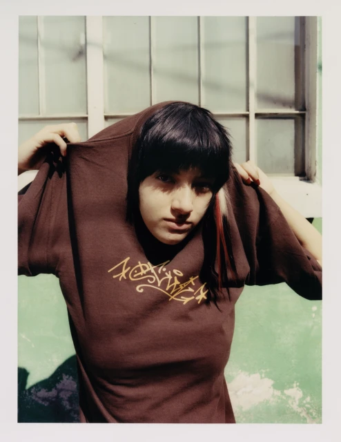 Portrait of a young woman with dark hair and a brown t-shirt with a graffiti logo posing indoors near a window and a light green wall.