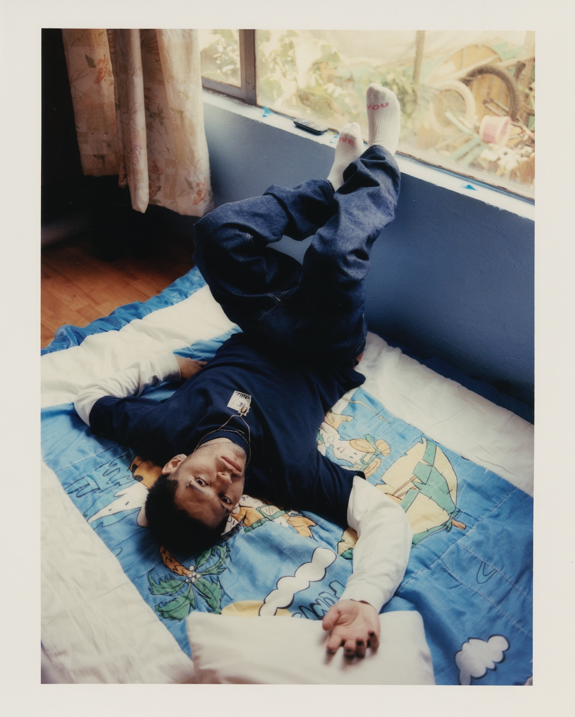Young man lying upside down on a bed with feet up against a window. The indoor shot captures his relaxed pose and casual attire.