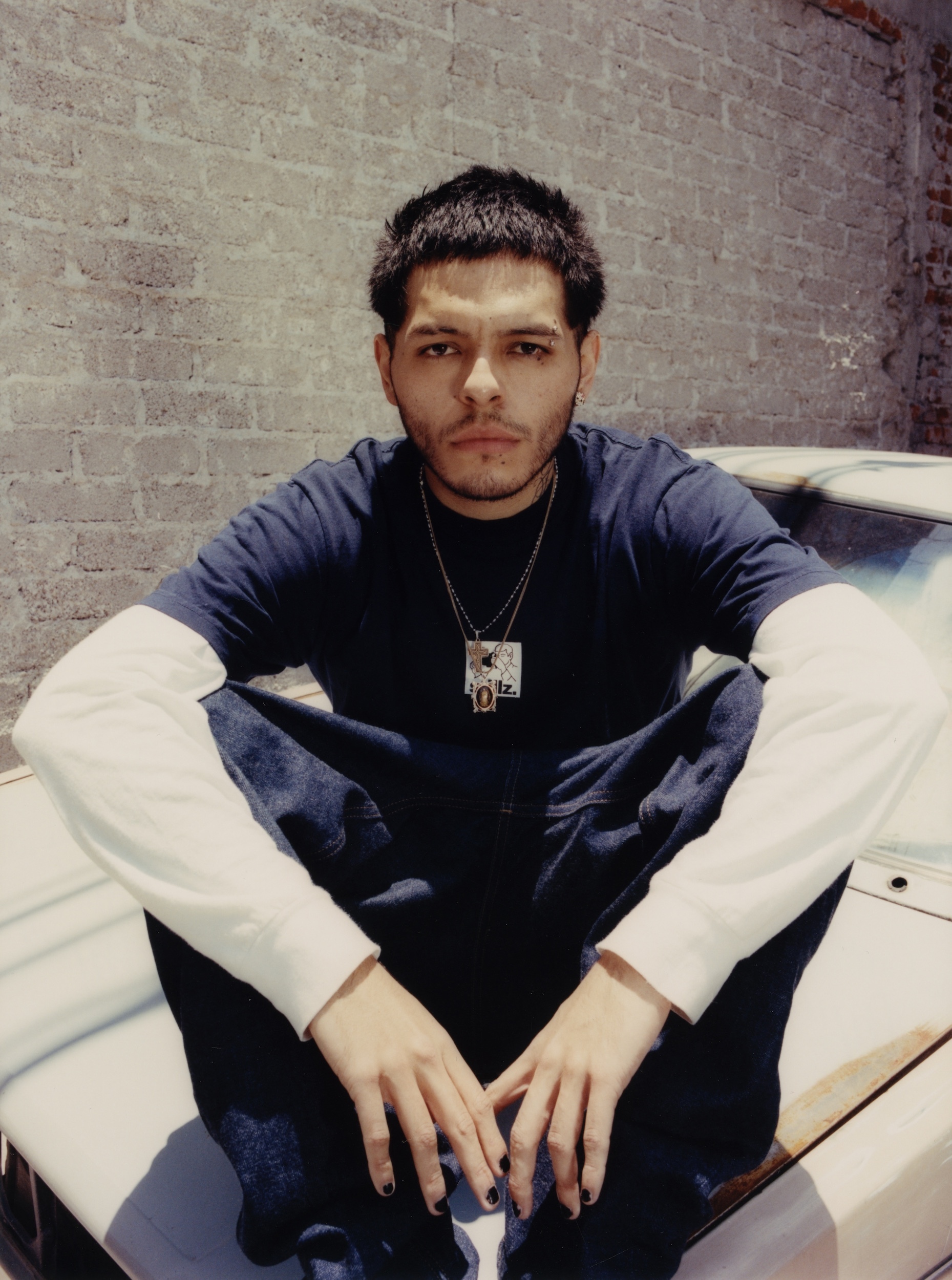 Portrait of a young adult male sitting on a car hood. He has dark hair and is wearing a dark shirt, light blue jeans, and a necklace.