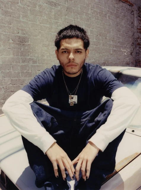 Portrait of a young adult male sitting on a car hood. He has dark hair and is wearing a dark shirt, light blue jeans, and a necklace.