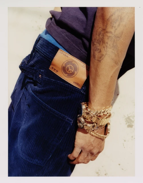 Close-up studio shot of a male model wearing blue corduroy pants and gold bracelets, showcasing a back pocket patch detail.