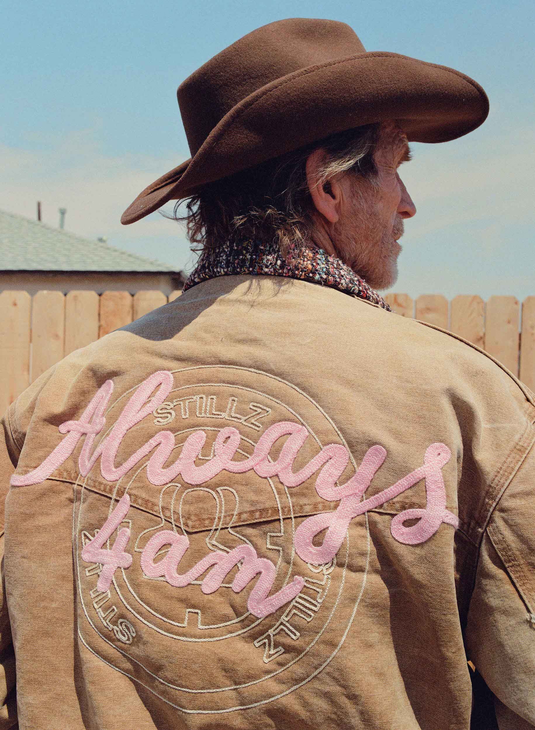 An adult male wearing a brown cowboy hat and jacket with an embroidered design, standing outdoors on a sunny day.