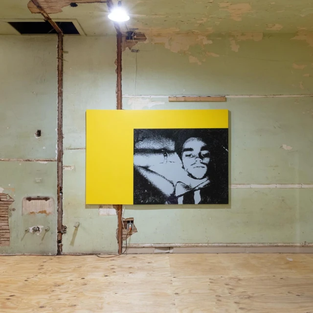 Portrait of a man on a black and white canvas hanging in a distressed room with a yellow square beside the canvas.
