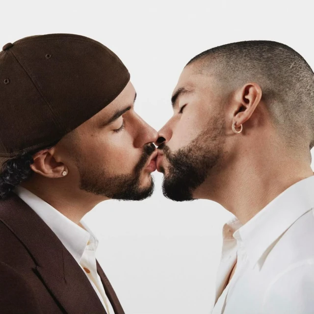 Two adult males in formal wear share a kiss against a light background, creating a portrait of intimacy and connection between them.
