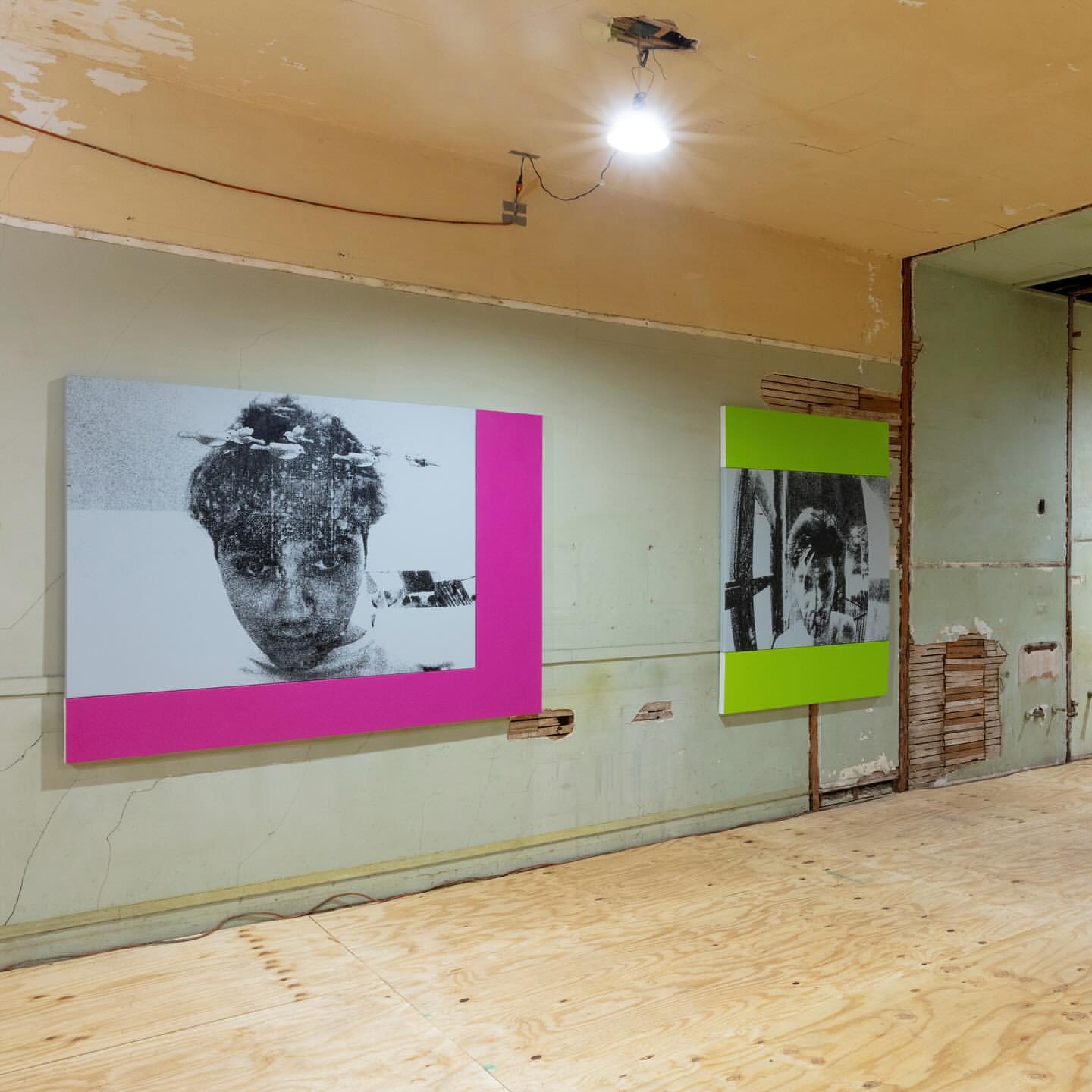 Interior view featuring black and white portrait artworks with magenta and lime green frames in a room with unfinished walls and floors.