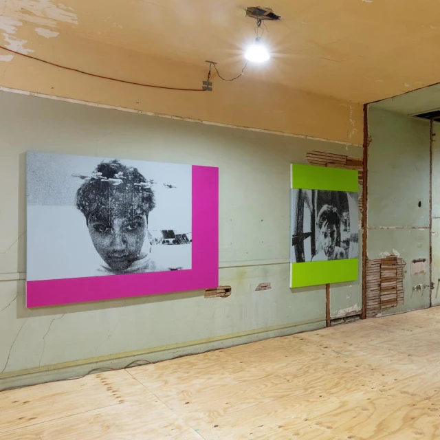 Interior view featuring black and white portrait artworks with magenta and lime green frames in a room with unfinished walls and floors.
