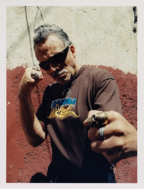A man with sunglasses and rings poses with fists up in front of a textured wall, wearing a t-shirt with a banana graphic.