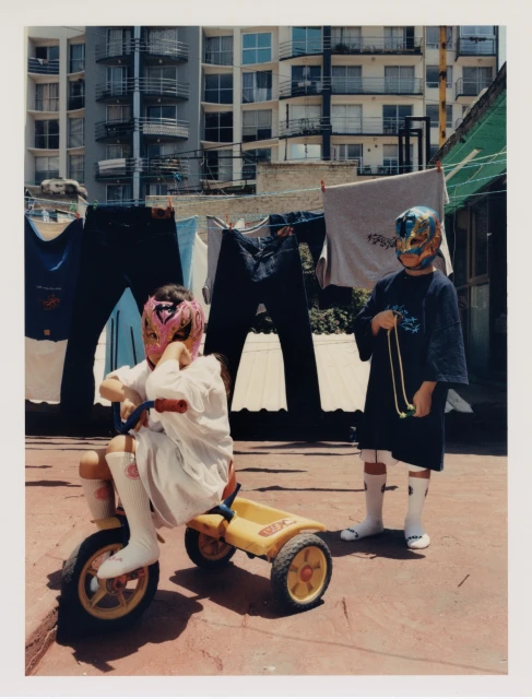 Two children in wrestling masks pose on a rooftop. One rides a yellow tricycle, with apartment buildings and hanging clothes in the background.