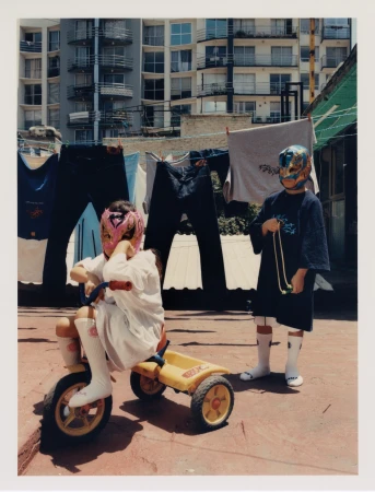 Two children in wrestling masks pose on a rooftop. One rides a yellow tricycle, with apartment buildings and hanging clothes in the background.