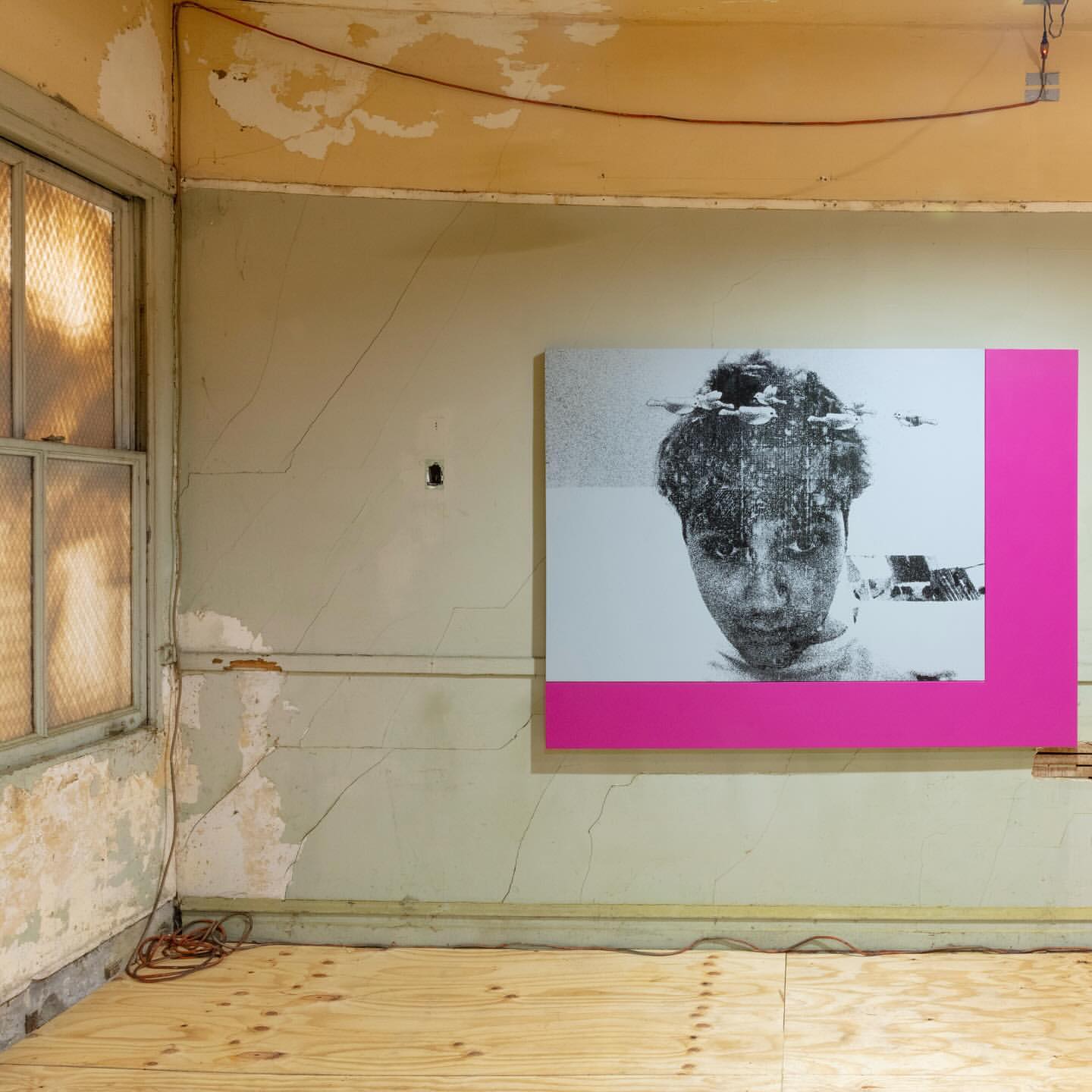 Photo of a black and white portrait with pink frame, hanging in a room with peeling walls, exposed wood floor, and a weathered window.