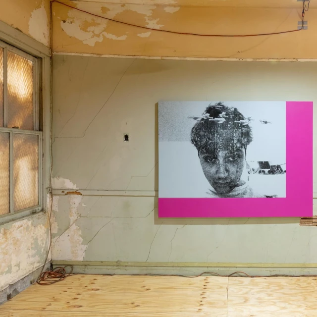Photo of a black and white portrait with pink frame, hanging in a room with peeling walls, exposed wood floor, and a weathered window.