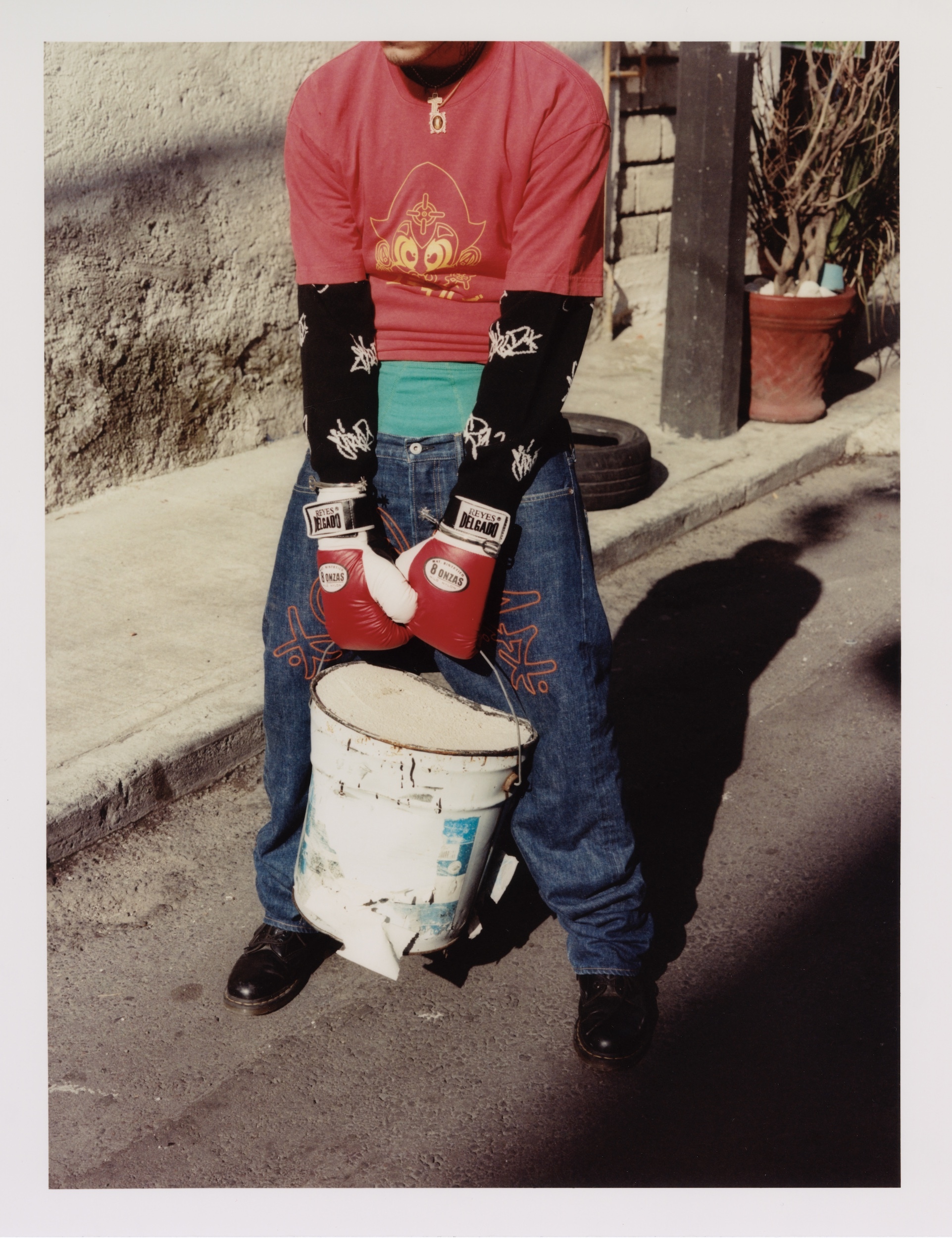 Person in boxing gloves carries a paint bucket in an urban environment, wearing casual clothing, captured in bright daylight.