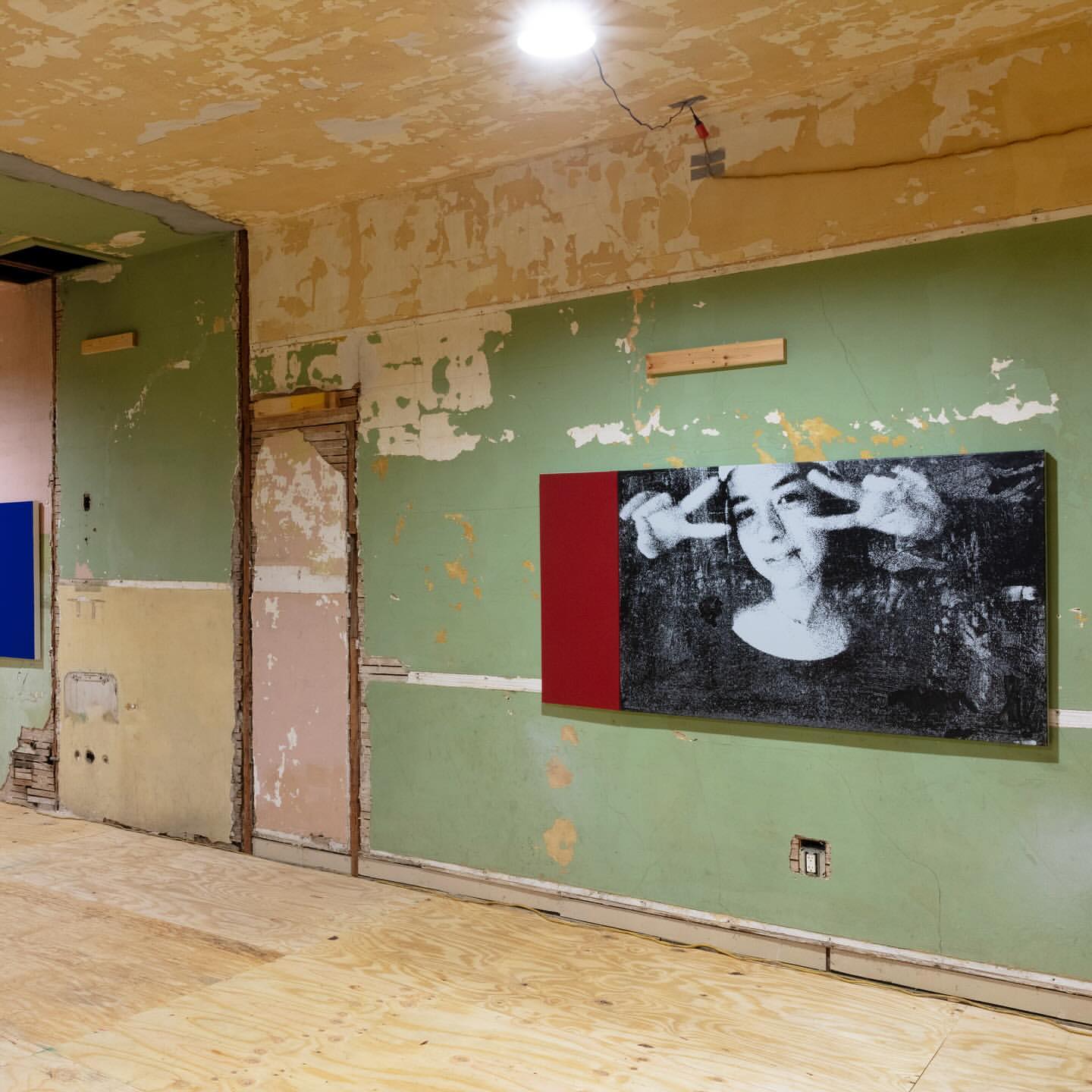 A black and white photograph of a person making a peace sign hangs on a green wall in a room under renovation with plywood floors.