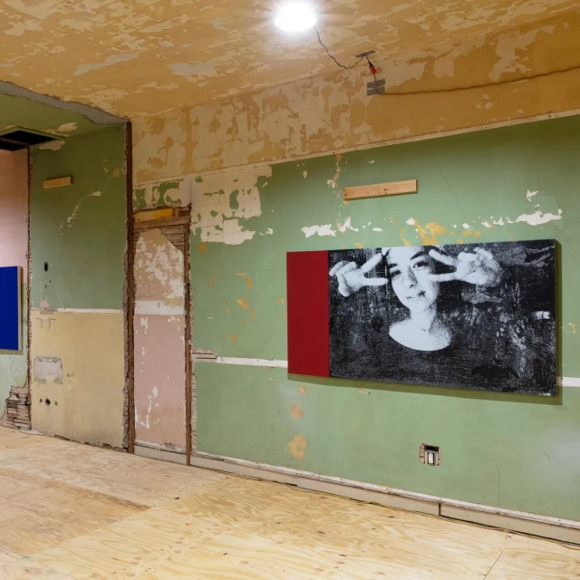 A black and white photograph of a person making a peace sign hangs on a green wall in a room under renovation with plywood floors.