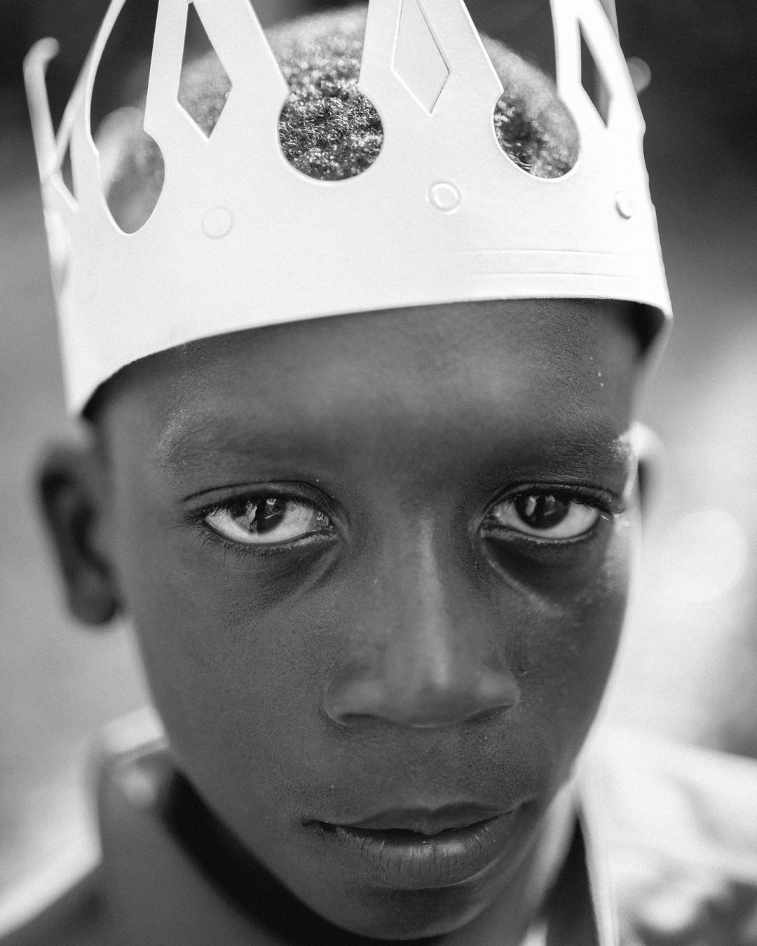 Portrait of a young African American boy wearing a white paper crown, photographed outdoors in black and white with shallow depth of field.