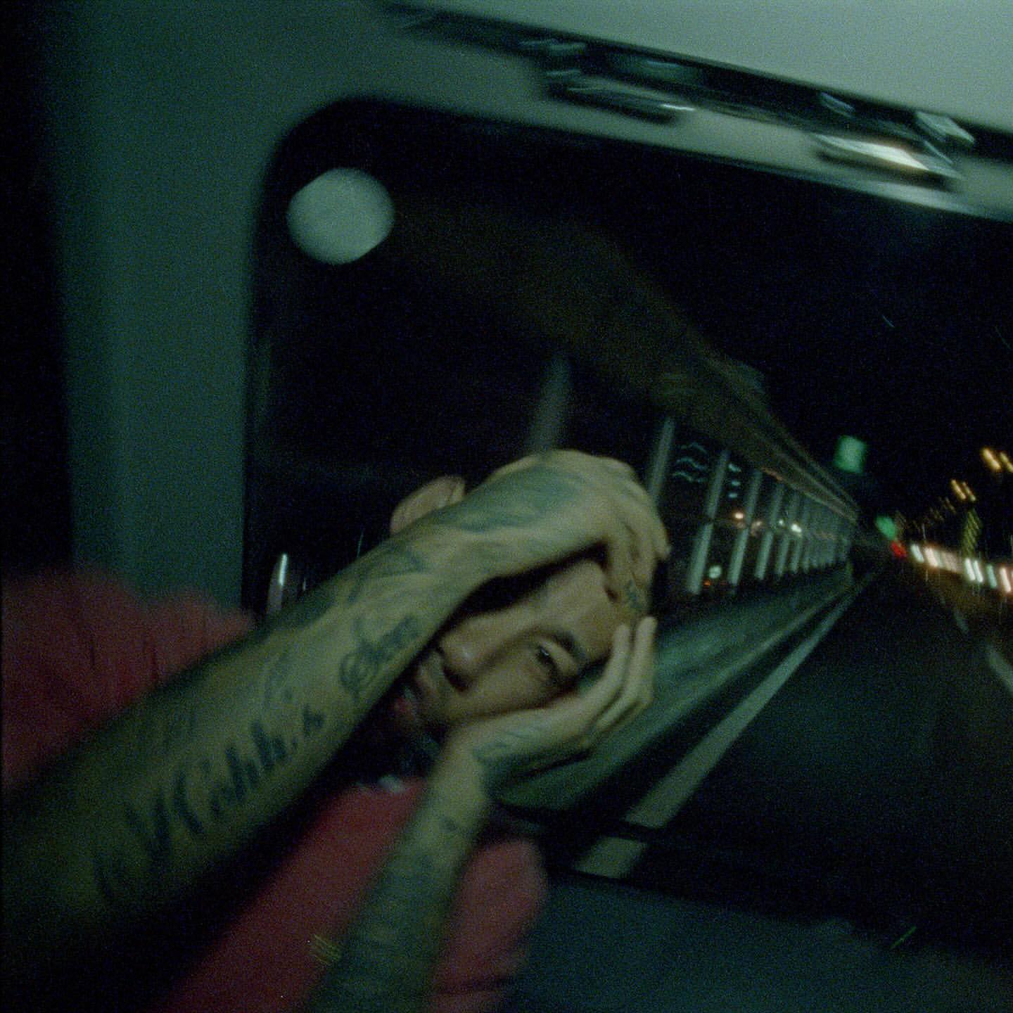 Adult male lying down, looking at camera, with tattoos on arms, inside vehicle at night with blurred city lights visible through window.