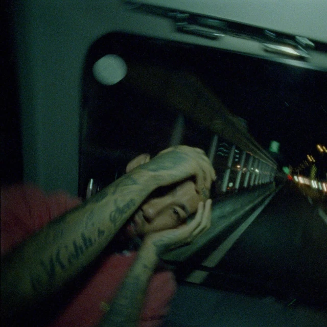 Adult male lying down, looking at camera, with tattoos on arms, inside vehicle at night with blurred city lights visible through window.