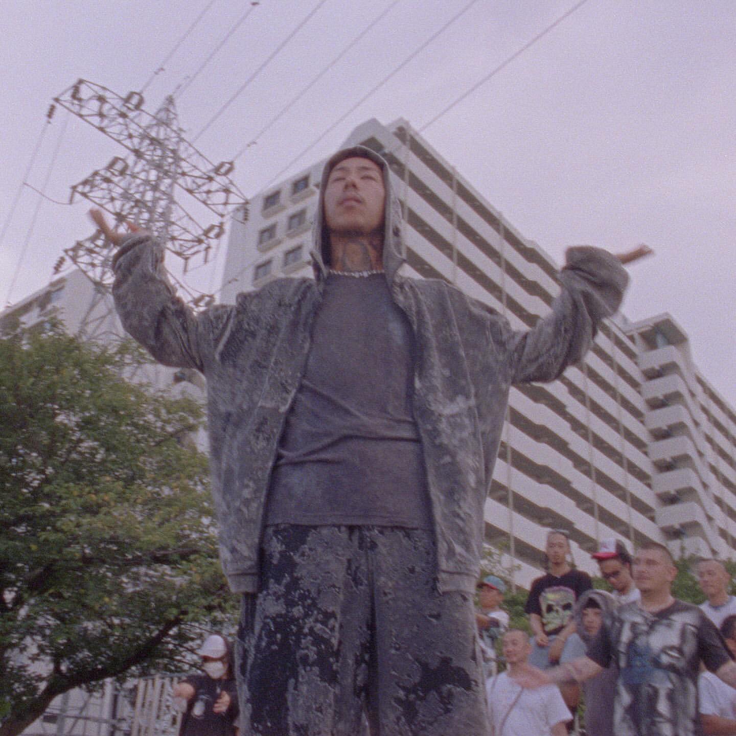 Asian male in hoodie stands with arms raised before a crowd and high-rise building, under overcast skies with powerlines overhead.