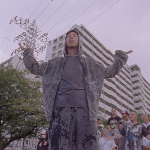 Asian male in hoodie stands with arms raised before a crowd and high-rise building, under overcast skies with powerlines overhead.