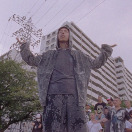 Asian male in hoodie stands with arms raised before a crowd and high-rise building, under overcast skies with powerlines overhead.