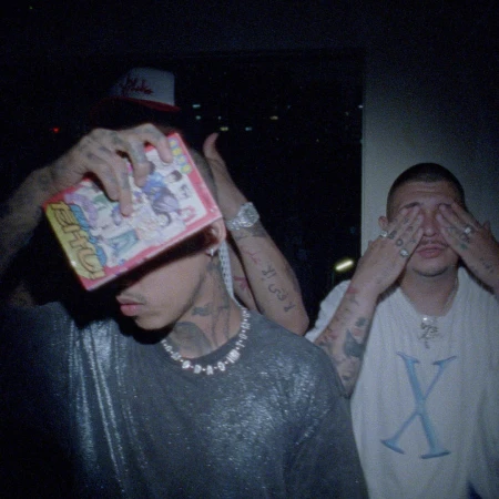 Two men in dimly lit setting with tattoos and necklaces, one holding a pink package to his face and the other covering his eyes.