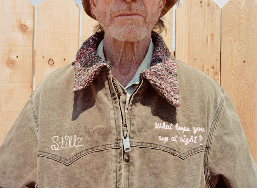 Close-up of an elderly man with a hat, wearing a brown work jacket with embroidered text against a light wood fence backdrop.
