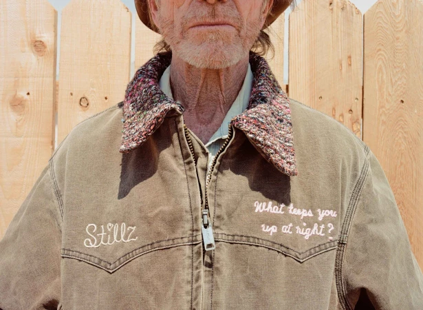 Close-up of an elderly man with a hat, wearing a brown work jacket with embroidered text against a light wood fence backdrop.