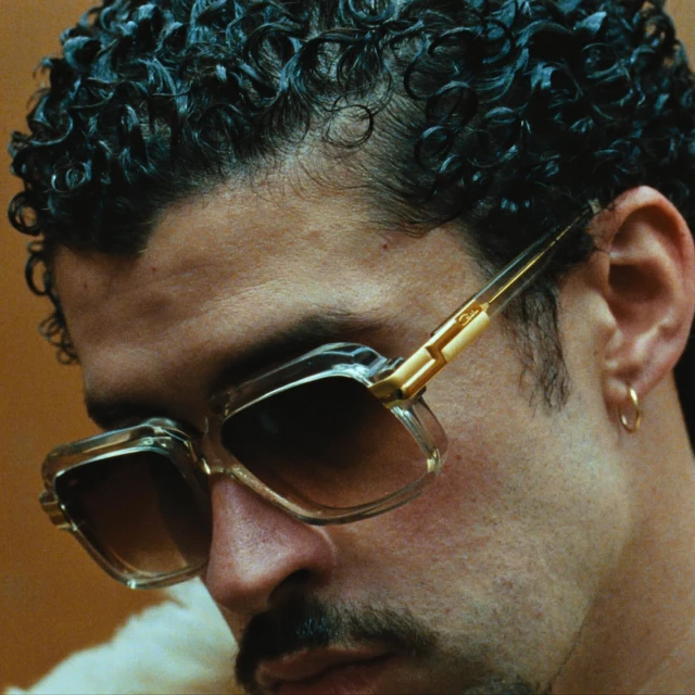 Headshot of a man with curly dark hair wearing clear-framed sunglasses and a gold earring, set against a tan background.