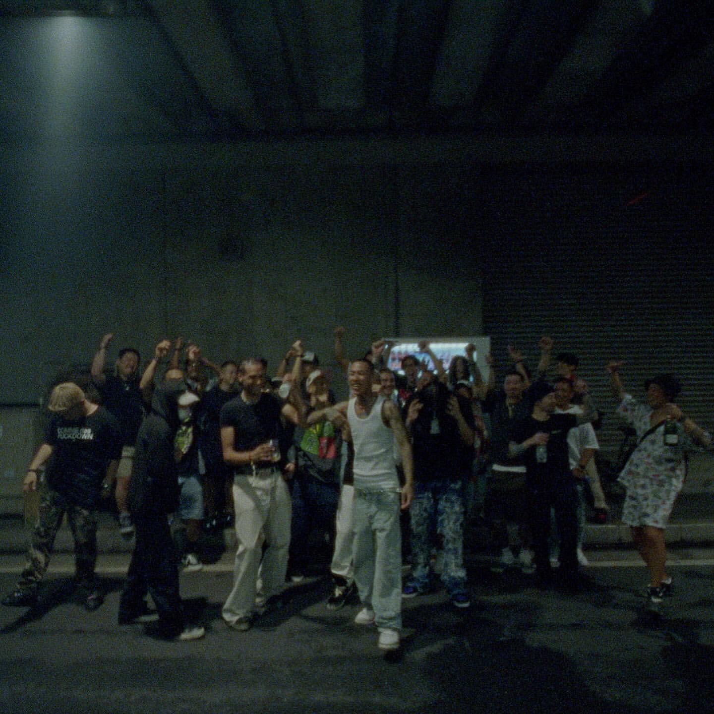 Large group of people gathers in an urban setting, laughing and celebrating with raised arms in a candid, low-lit scene.