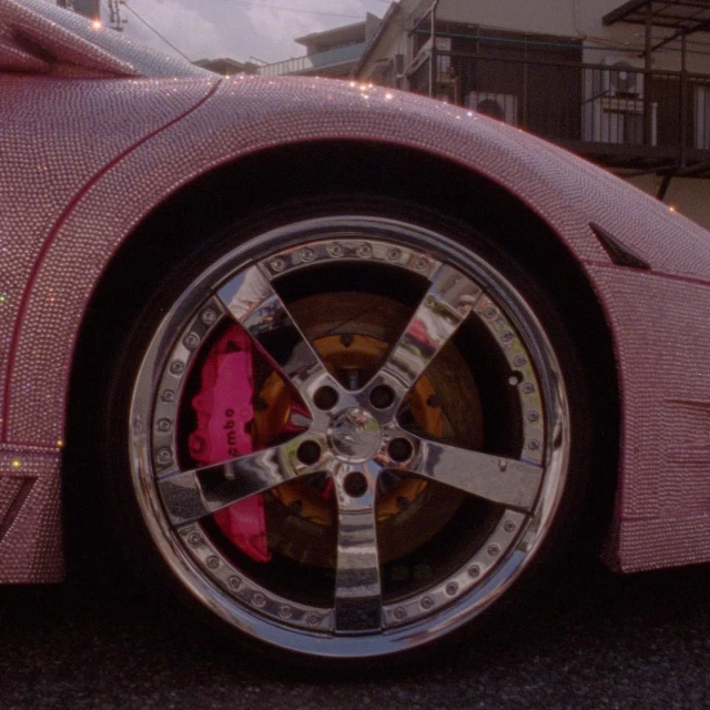 Close-up of a pink car encrusted with crystals, highlighting the chrome rim and pink brake caliper. The vehicle is outdoors.