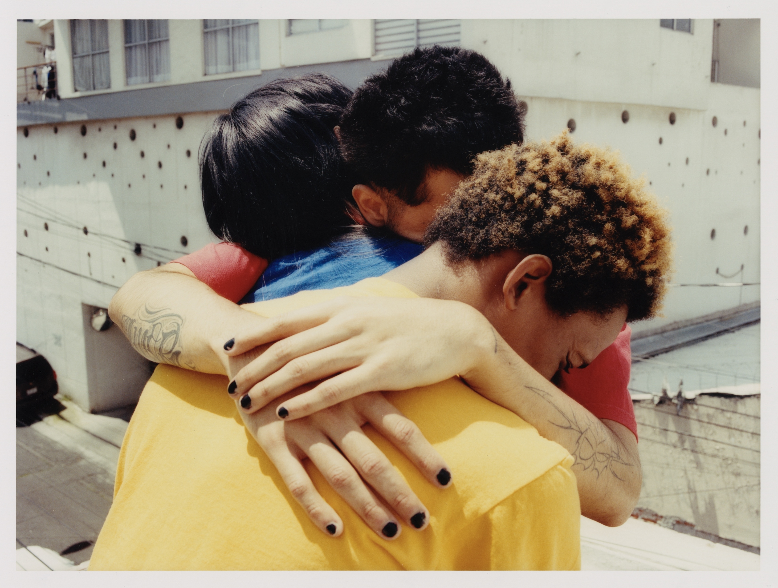 Three people embracing outdoors in a supportive group hug, showcasing close connection, against an urban building backdrop on a sunny day.