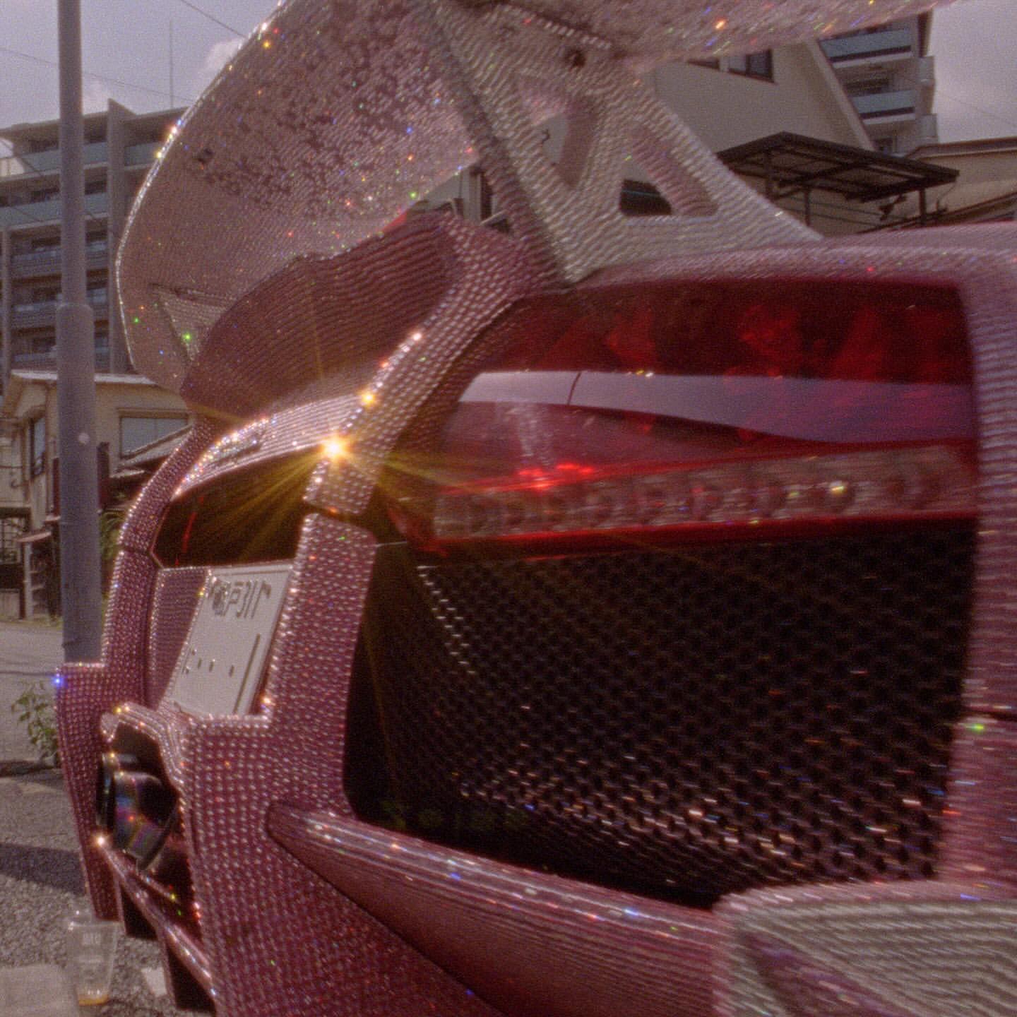 Rear view of a pink car covered in rhinestones, featuring a large spoiler. The vehicle is parked on a street with buildings visible.
