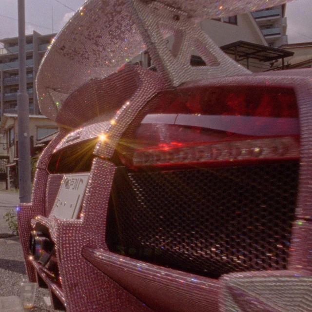 Rear view of a pink car covered in rhinestones, featuring a large spoiler. The vehicle is parked on a street with buildings visible.