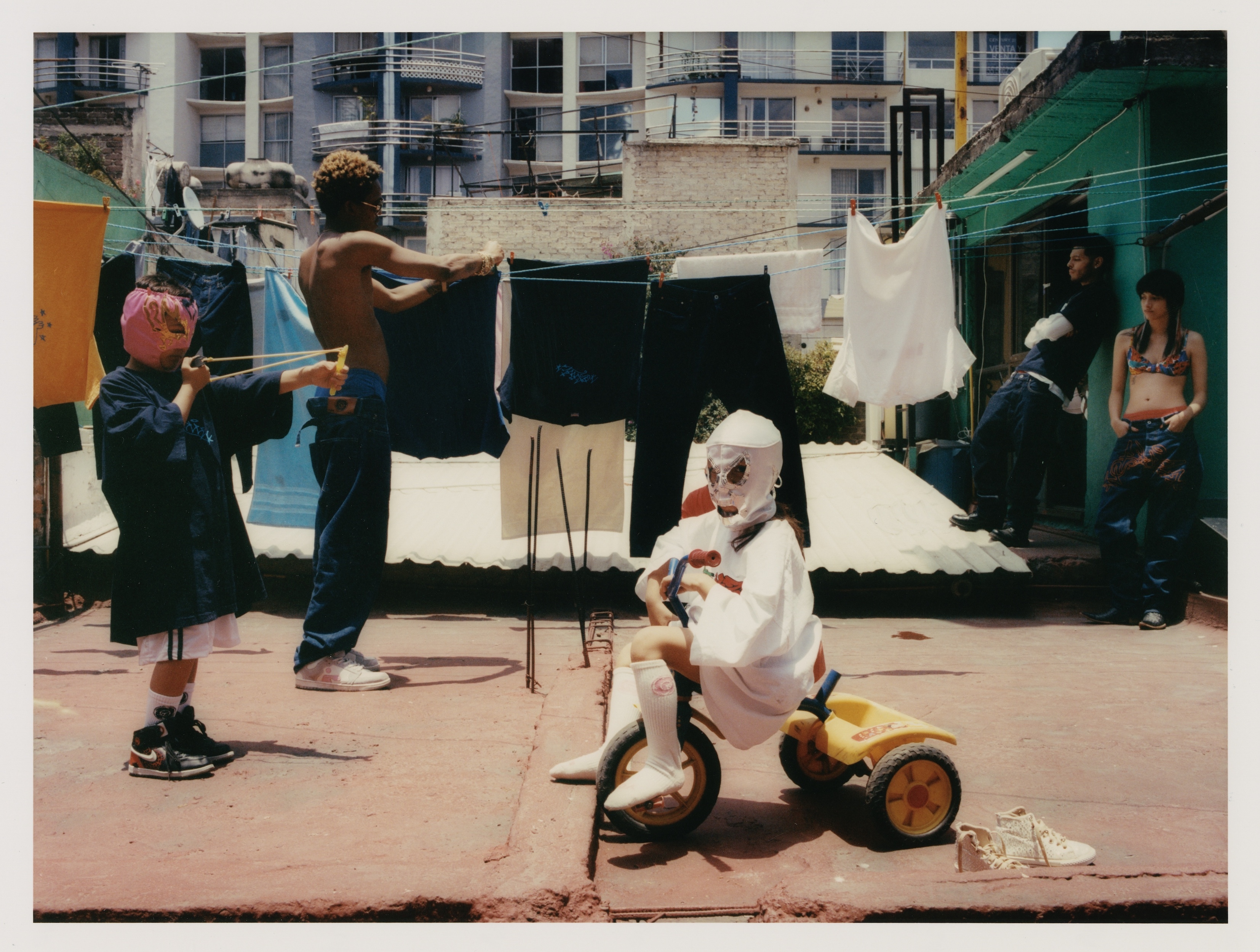 Several figures gather on a rooftop clothesline scene, one in a mask on a yellow tricycle, another shooting a toy gun, with buildings in the distance.