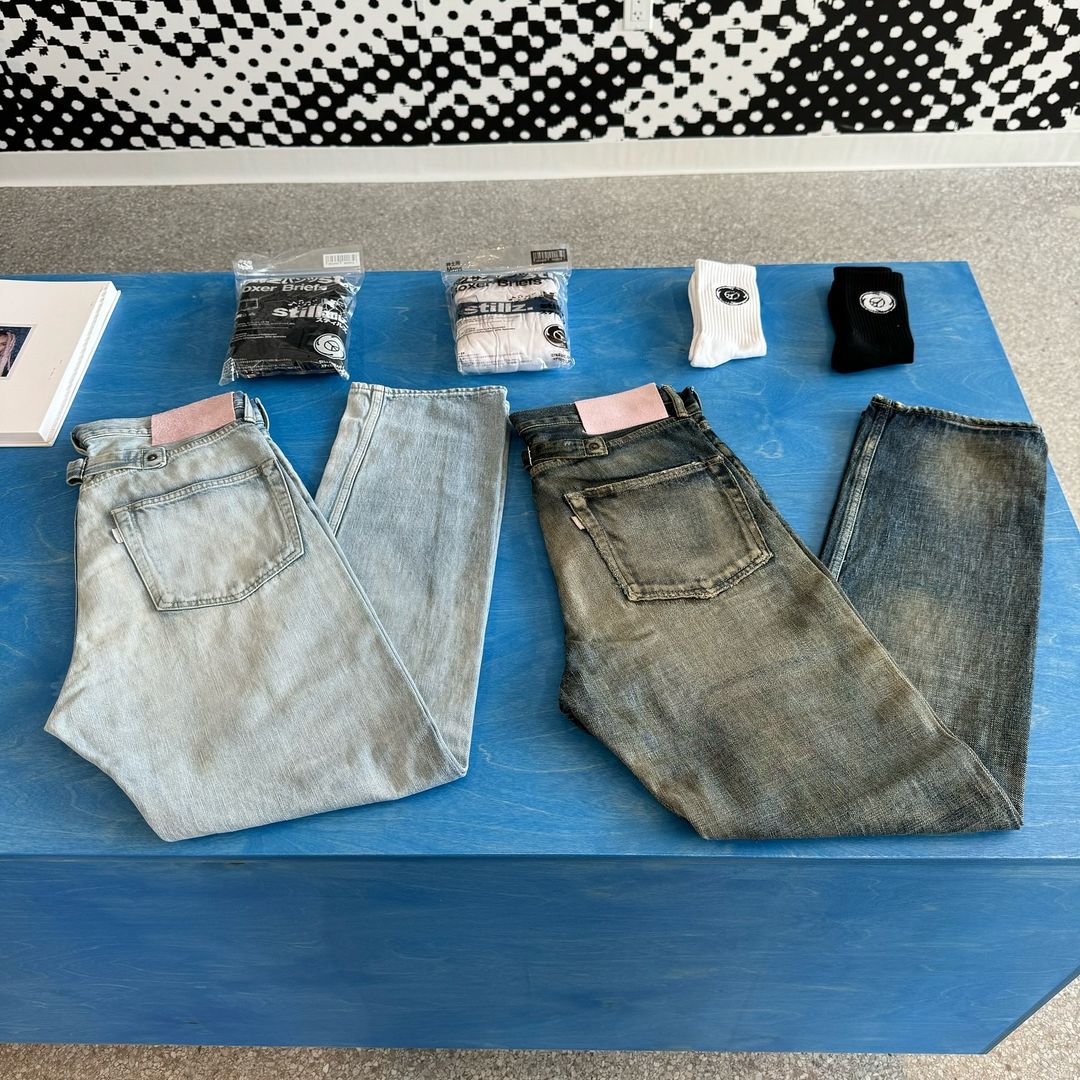 Two pairs of jeans are displayed on a blue surface, accompanied by packaged socks and a patterned backdrop in an indoor setting.