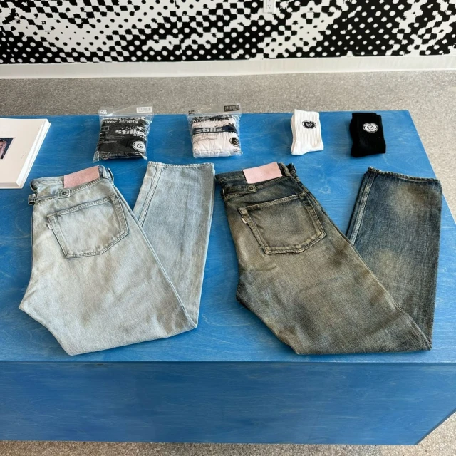 Two pairs of jeans are displayed on a blue surface, accompanied by packaged socks and a patterned backdrop in an indoor setting.
