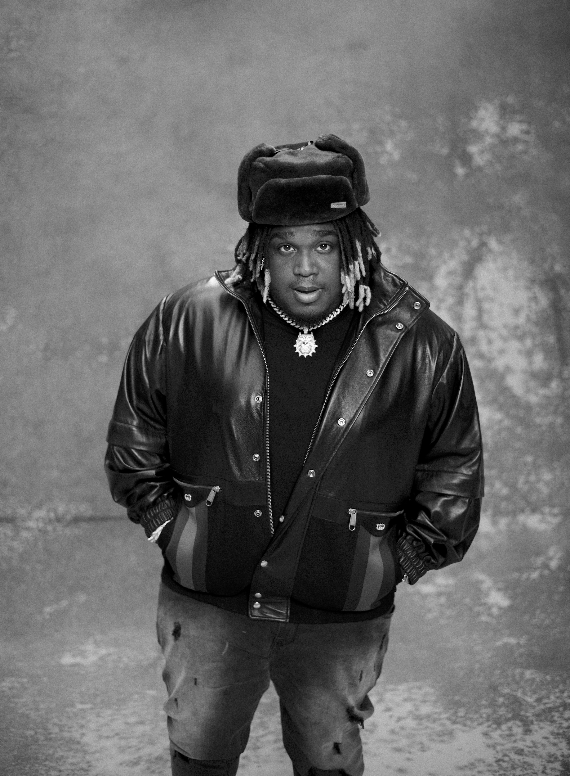 Black and white portrait of an adult male with dreadlocks wearing a trapper hat, leather jacket, and ripped jeans on a mottled background.