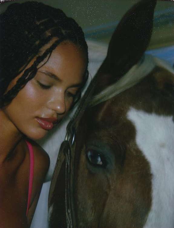 A woman with dark braided hair rests her head against the side of a brown and white horse, close-up shot with a shallow depth of field.