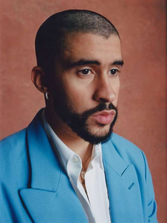 Portrait of adult male with a beard wearing a light blue suit jacket and white collared shirt, against a light brown background.
